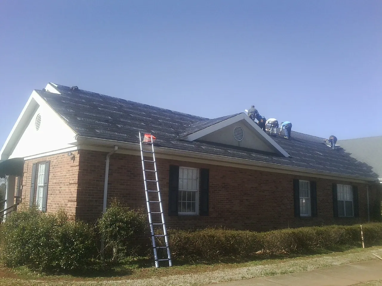 Crew tearing shingles off a brick ranch roof with a ladder set — Emergency Roof Repair work by our crew in Lore City