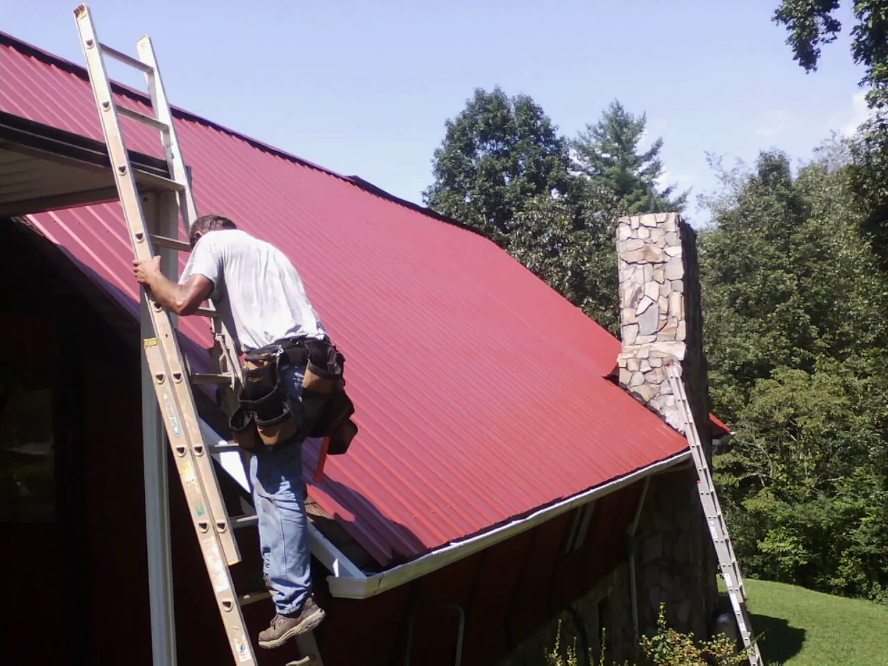 Roofer on a ladder beside a red metal roof and stone chimney — Soffit & Fascia Repair by Roofing Brothers in Lore City